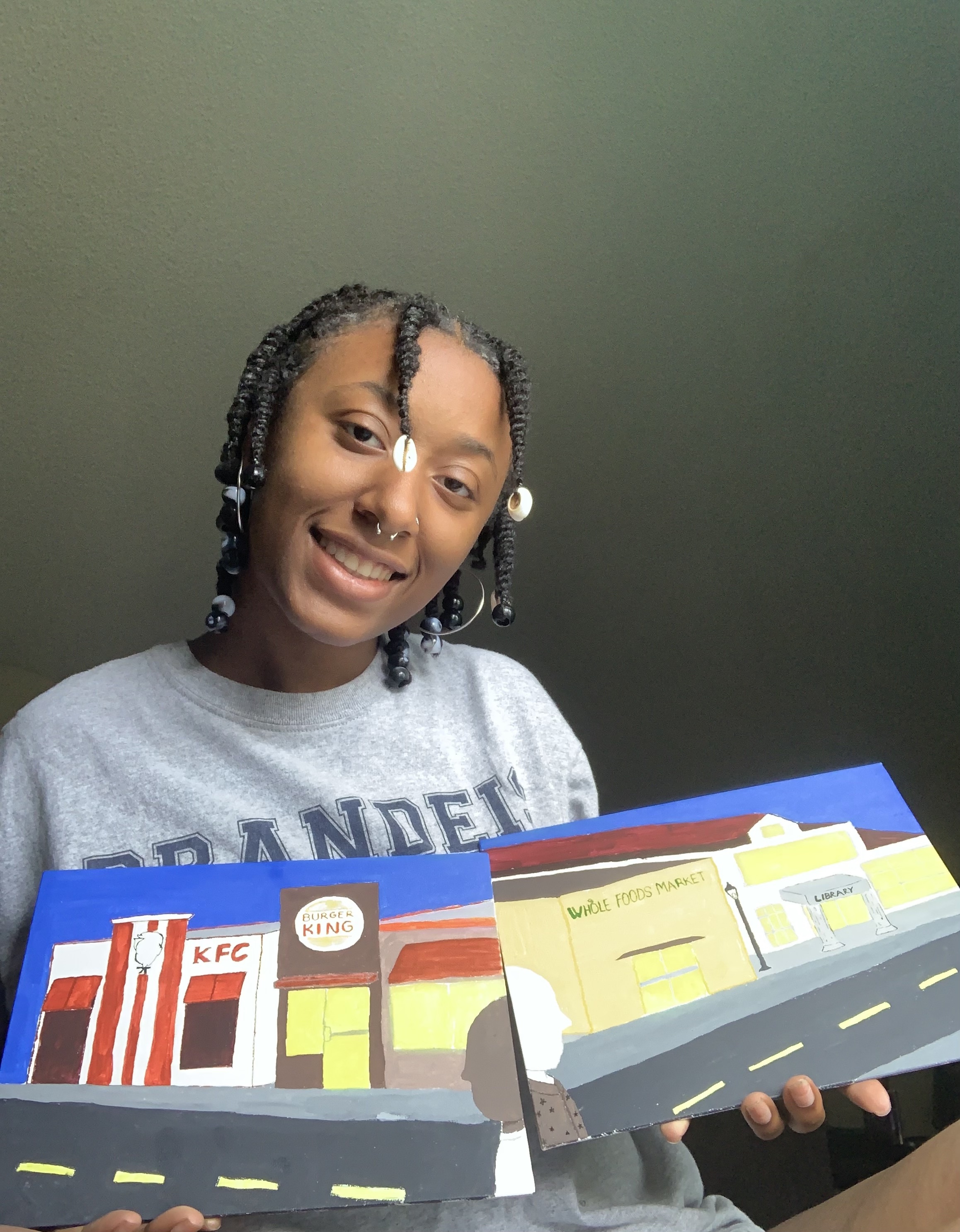 Image of young Black woman with seashells and beads at ends of braids holding two colorful painted canvases